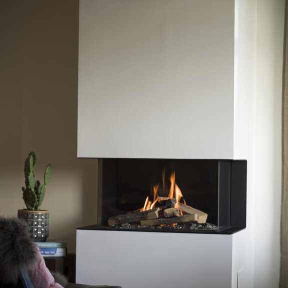 Gas-Kamin VISIO 90 3S als dreiseitiger Kamin mit ruhiger Gasflamme in einer weissen Verkleidung neben Pflanze und Lesesessel
